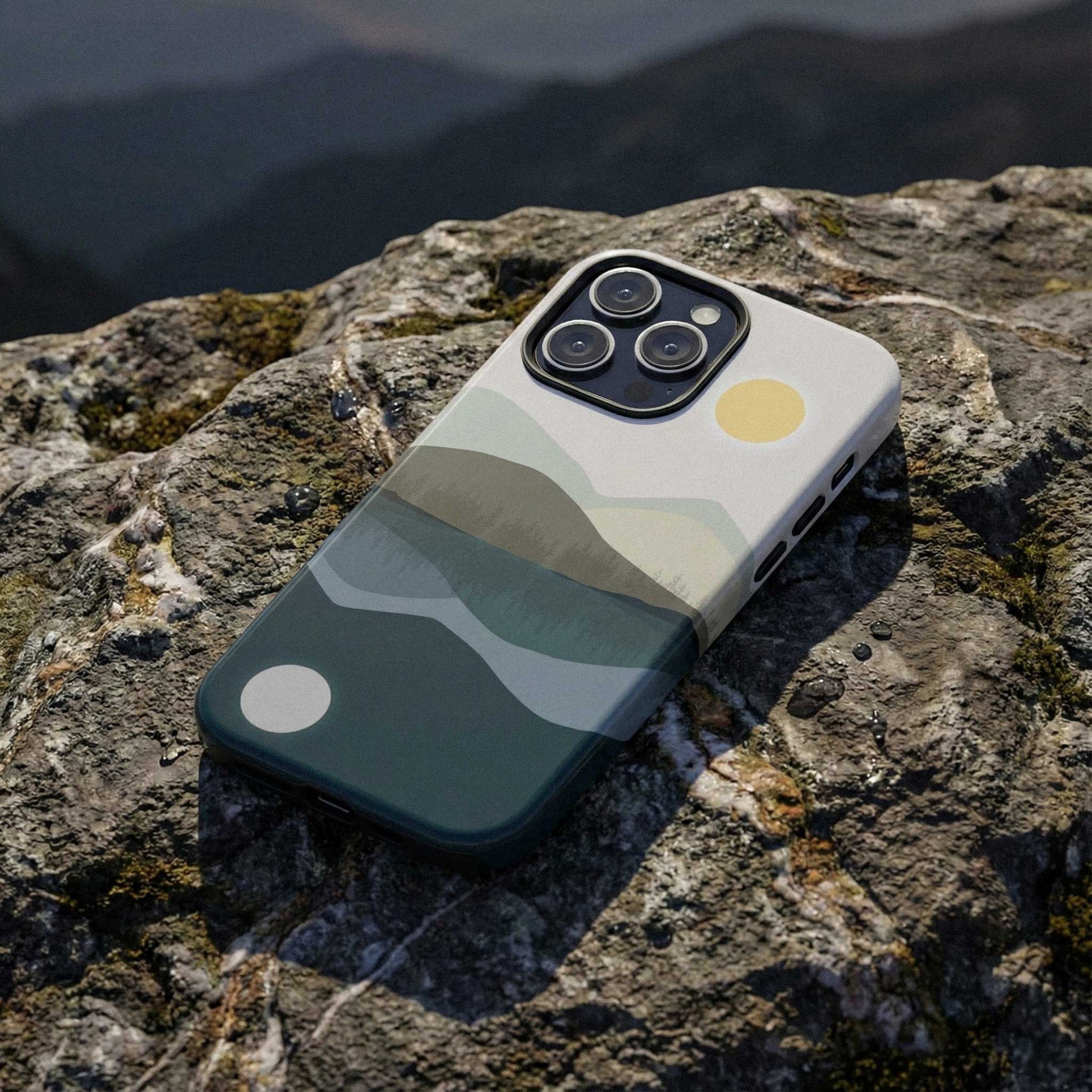 Phone case with an abstract mountain and lake reflection illustration, displayed on a rock against, designed for iPhone and Samsung models.