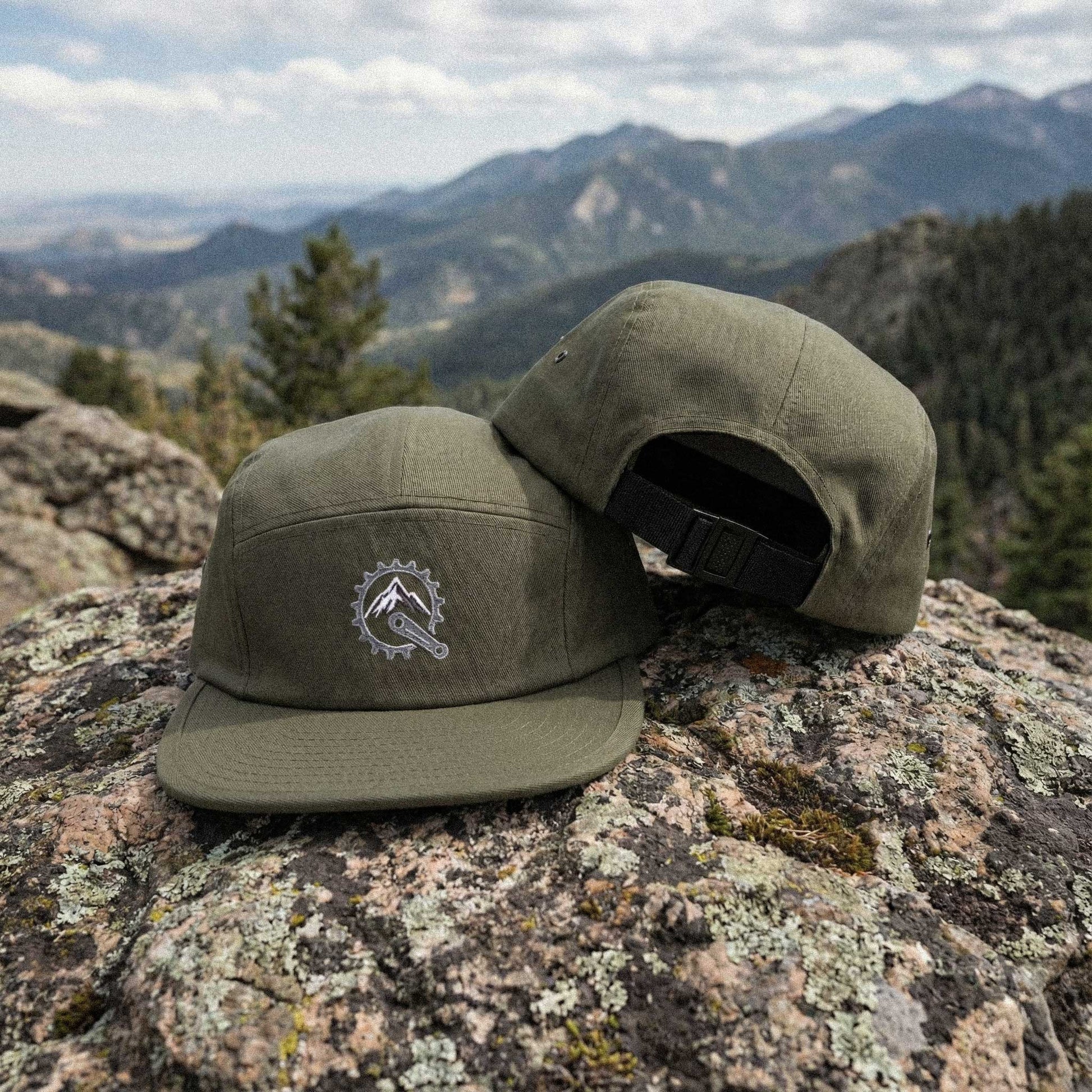 Two olive green panel caps with embroidered Mountain Bike Sprocket / Mountain Peak graphic on a rocky surface with mountains in the background by Kempain Family Outfitters.