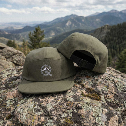 Two olive green panel caps with embroidered Mountain Bike Sprocket / Mountain Peak graphic on a rocky surface with mountains in the background by Kempain Family Outfitters.
