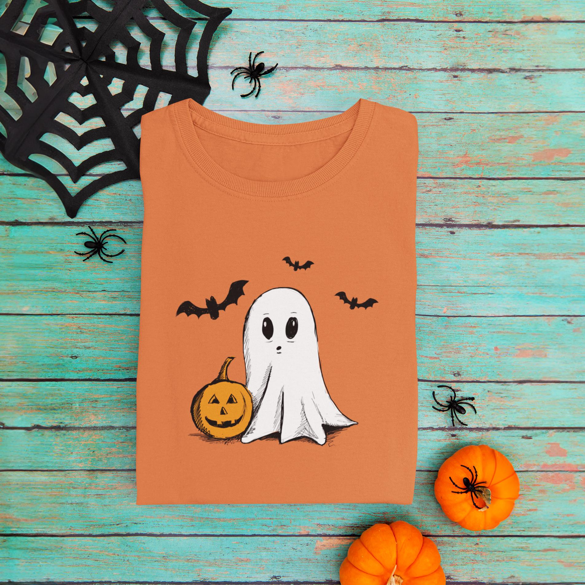 Orange Halloween shirt folded on a rustic wooden background, surrounded by Halloween items like plastic spiders, a spider web, and a pumpkin, creating a festive spooky atmosphere.