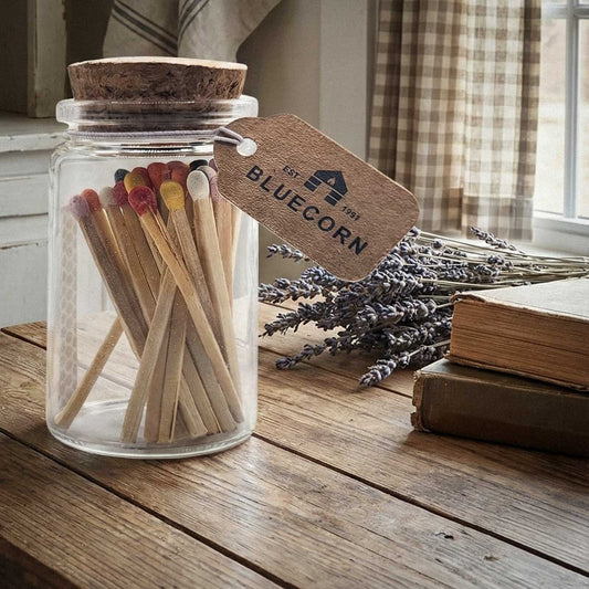 Jar of matches with a 'Bluecorn Candles Logot' tag on a wooden surface with lavender and books in the background.
