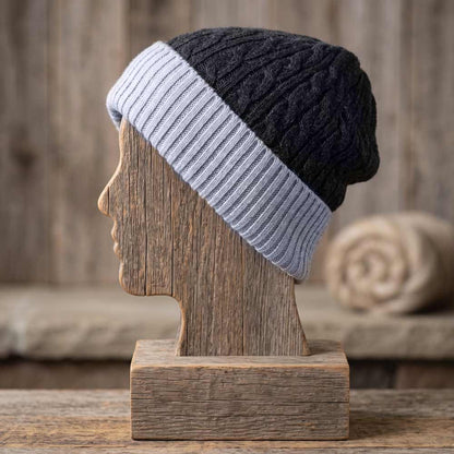 A two-tone cable-knit beanie hat displayed on a wooden mannequin head stand. The hat features a black upper section and a light gray ribbed brim, set against a rustic wooden background.