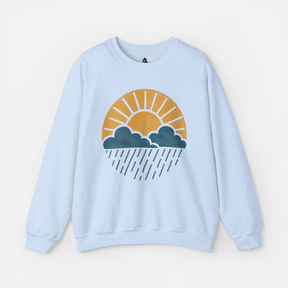 Flat lay of Sun Over Clouds Minimalist Sweatshirt in light blue fabric on a white background, featuring a simple line art illustration of a sunrise over clouds and rain within a subtle circle, printed on a classic unisex crewneck.