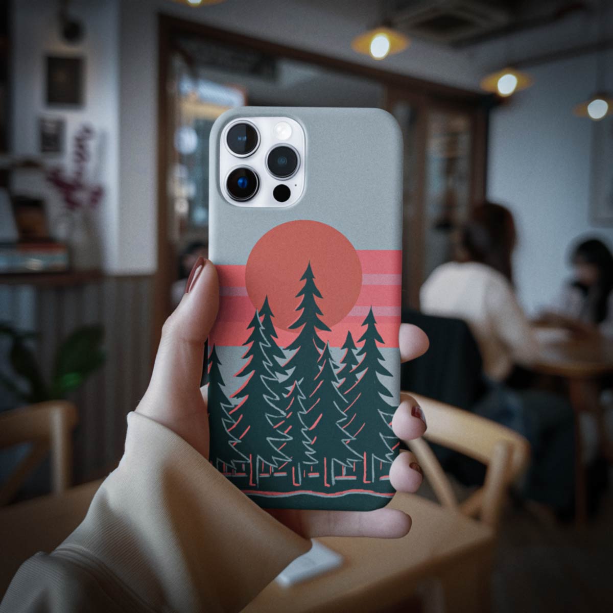 A hand holds a phone case featuring a retro sunset landscape design with pine trees, a large orange sun, and pink stripes on a gray phone, set against a blurred cafe background.