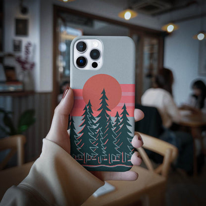 A hand holds a phone case featuring a retro sunset landscape design with pine trees, a large orange sun, and pink stripes on a gray phone, set against a blurred cafe background.