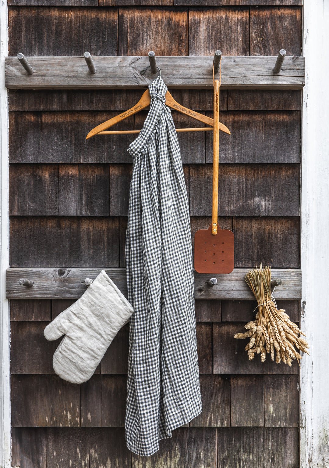 The Leather Huntsman Flyswatter displayed on a wooden hook on rustic cedar siding to showcase the farmhouse appeal.