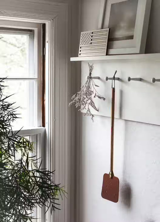 The Leather Huntsman Flyswatter by Farmhouse Pottery sold by Kempain Family Outfitters displayed on a hook in farmhouse setting.