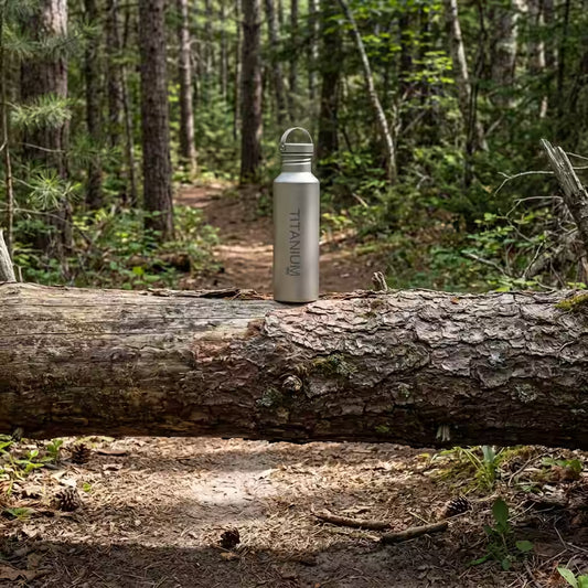 A VARGO titanium water bottle with 'TITANIUM' printed vertically, sitting on a fallen log in a forest setting.