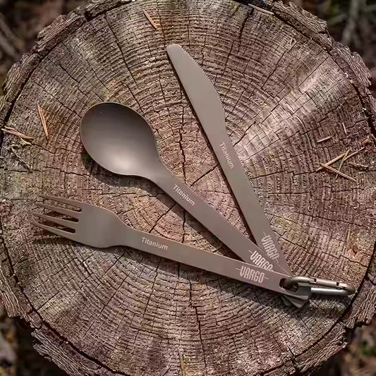 A titanium camping utensil set with a spoon, knife, and fork attached to a carabiner, arranged on a weathered tree stump outdoors.