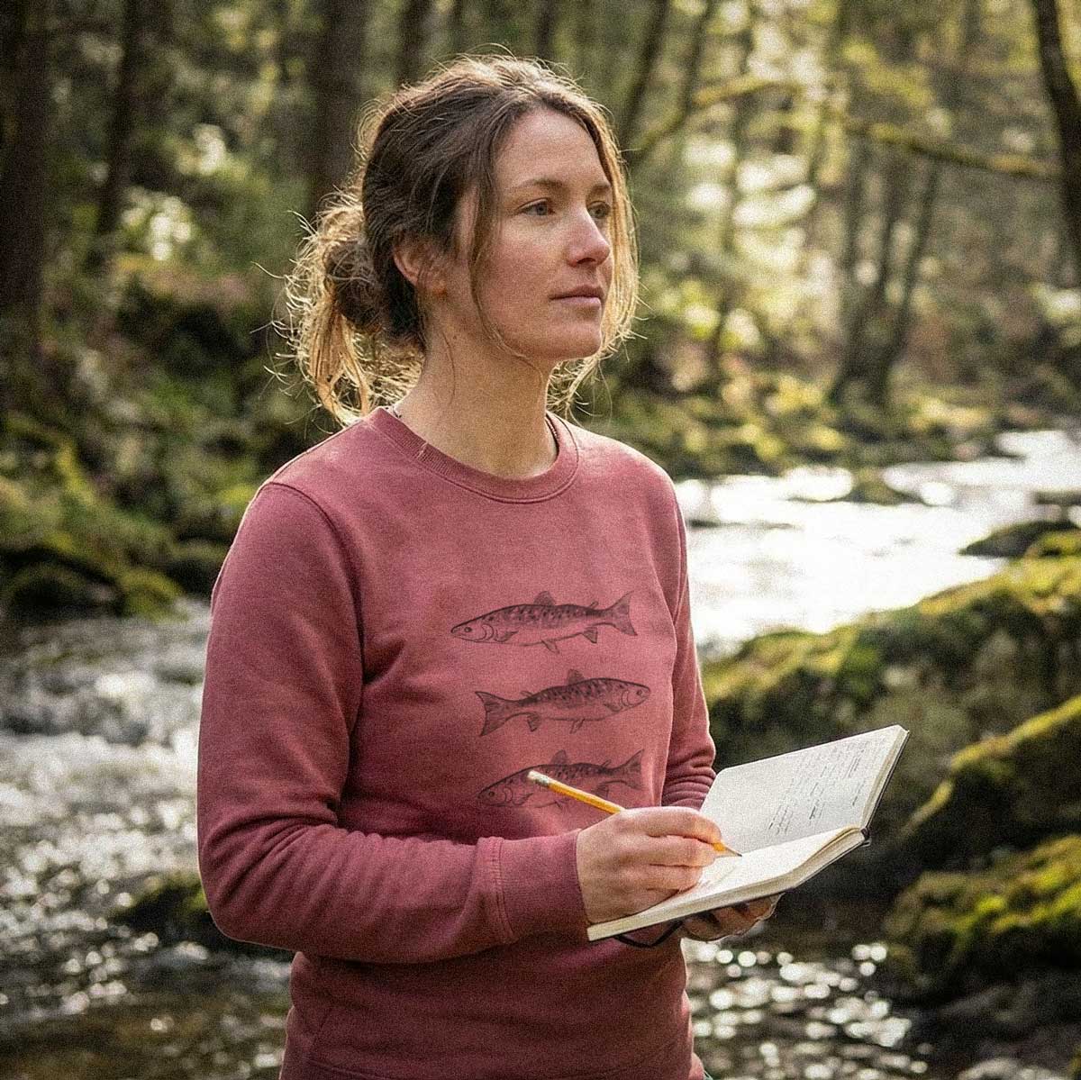 Female model wearing the Crimson Triple Fish Artist Crewneck in a forest setting with a field sketchbook. This 100% ring-spun cotton, 6.4 oz sweatshirt features original hand-drawn fish illustrations by Jordan Kempain.