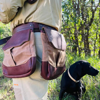 A person outdoors wearing a brown and tan leather-and-canvas waist bag system with two large pouches, with a black hunting dog sitting nearby in a grassy wooded area.