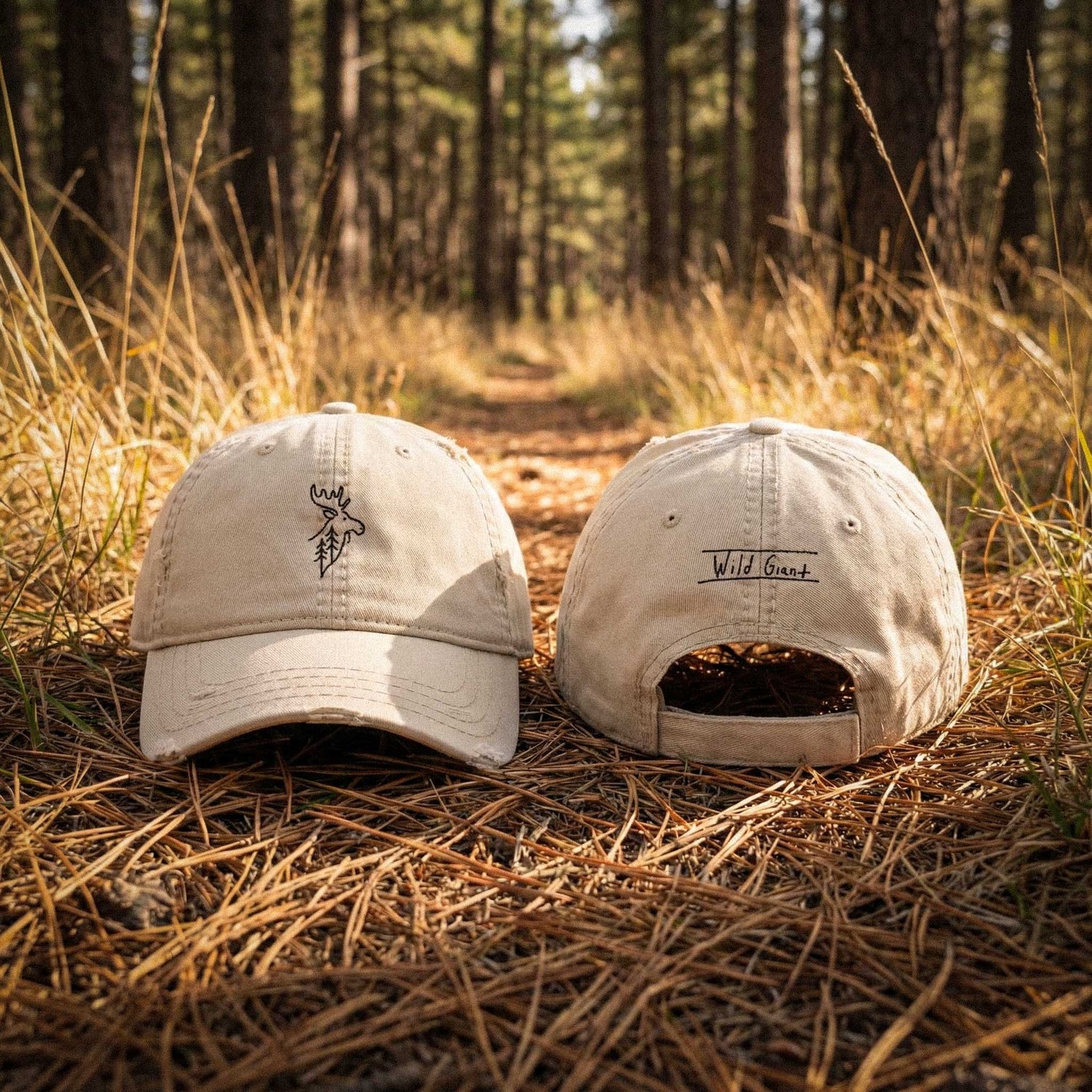 Two beige caps with wild moose embroidery and "wild giant" hand drawn text on back on a forest floor