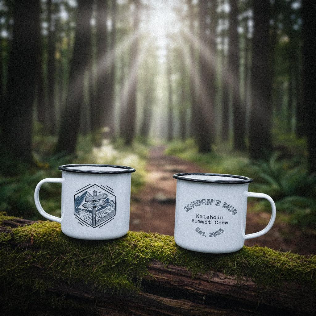 Two personalized camping mugs with trail sign design on a mossy log in a deep forest path.